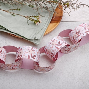 May include: A decorative paper chain featuring a repeating floral pattern in shades of dusty pink, mauve, and white. The chain consists of interlocking paper loops, creating a continuous garland design with a soft, matte finish on the paper surface.