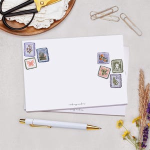 May include: White stationery featuring decorative postage stamp illustrations. A white pen with gold detailing, gold paper clips, and scissors are visible. Dried flowers and greenery are arranged to the side.