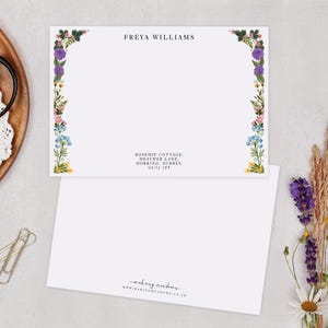 a blank card with a floral border, placed on a table with various items such as a pair of scissors, a white lace doily, and some dried flowers.