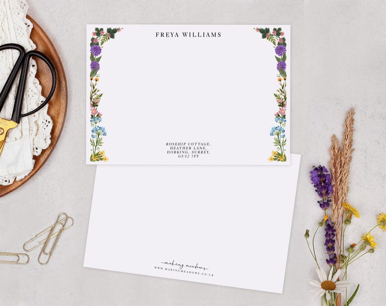 a blank card with a floral border, placed on a table with various items such as a pair of scissors, a white lace doily, and some dried flowers.