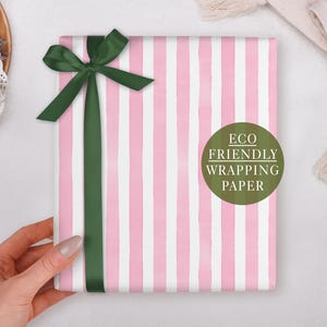 May include: A gift wrapped in pink and white striped paper, adorned with a dark green ribbon and bow. A green circle with the text "Eco Friendly Wrapping Paper" is visible. The gift is held by a hand.