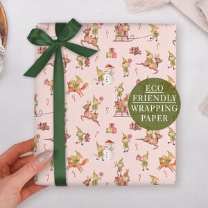 A hand holding a pink gift box with a green ribbon, decorated with a festive pattern of Christmas-themed characters and presents.