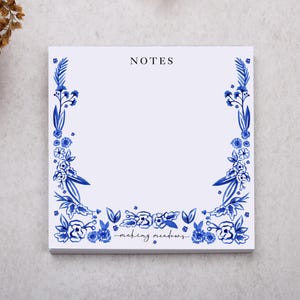 May include: Square white notepad with blue floral border and the word "NOTES" at the top. The bottom of the notepad has the words "making meadows" in script. The notepad is on a light grey surface.
