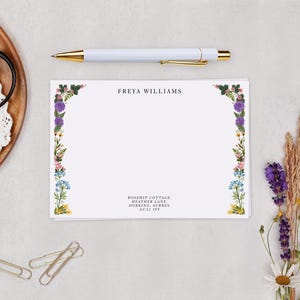 a blank card with a floral border, a pen, a gold pen, a pair of scissors, a wooden plate with a lace doily, and some dried flowers.