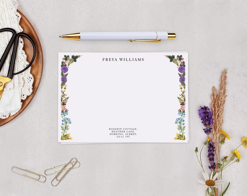a blank card with a floral border, a pen, a gold pen, a pair of scissors, a wooden plate with a lace doily, and some dried flowers.