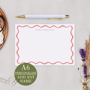 May include: A white A6 personalised stationery card with a wavy pink border and the name "ROBYN BRECKLE" printed in the upper right corner. A white pen with gold accents and a green circle with the text "A6 PERSONALISE ADD ANY NAME" are also visible.