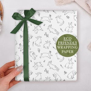 A hand holding a gift wrapped in a white paper with a green ribbon and a green circle with text that reads "ECO FRIENDLY WRAPPING PAPER".