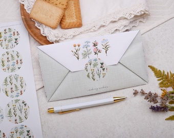 May include: An envelope with a floral design and a matching sticker sheet. The envelope is light green with a white flap featuring watercolor flowers. A white pen with gold accents and cookies are also present.