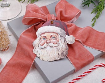 Peut inclure: Ornement de Père Noël avec un bonnet rouge et une barbe blanche posé sur un coffret gris, orné d'un ruban de velours rose poudré. Un petit sapin de Noël et une canne en sucre sont également visibles, créant une scène de fête.