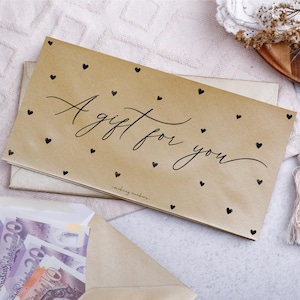 Peut inclure: Une enveloppe cadeau marron clair avec l'inscription "A gift for you" en écriture cursive, ornée de petits cœurs noirs. L'enveloppe est posée sur une autre enveloppe. Une troisième enveloppe ouverte révèle de l'argent.