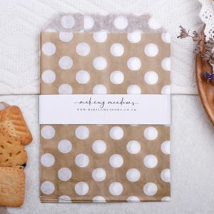 May include: Brown paper bags with white polka dots, wrapped with a white band featuring the text "making meadows" and a website address. A biscuit is partially visible in one bag. The image is set on a white surface.