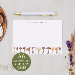 May include: A6 personalised stationery featuring a watercolour mushroom design. The stationery has the text "MR & MRS CHELSEA" at the top and a row of colourful mushrooms along the bottom. A white pen with gold accents rests above the stationery.