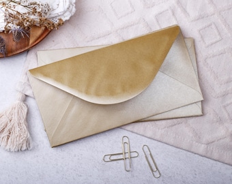 May include: Three kraft paper envelopes with a gold flap, and two gold paper clips. The envelopes are on a textured, pale pink surface, with a decorative tassel and a wooden tray with dried flowers in the background.