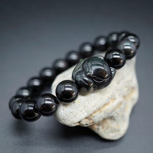 May include: A black beaded bracelet with a carved focal bead. The bracelet is made of round, polished black beads. The focal bead is a carved figure. The bracelet is resting on a light gray stone.