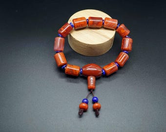 May include: A handcrafted bracelet with reddish-orange, cylindrical beads strung on a blue cord. The bracelet has a central, larger bead with a dangling tassel of smaller beads. The beads are displayed on a wooden stand against a dark background.