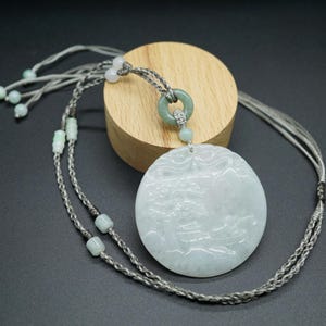 May include: A circular pale green jade pendant featuring a carved landscape design. The pendant is suspended from a grey cord necklace with small, light green beads. The necklace is displayed on a wooden stand against a dark background.