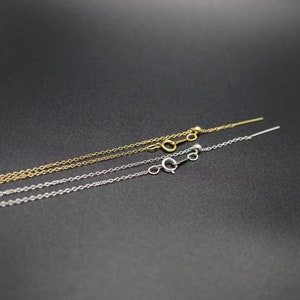 May include: Two delicate chains, one gold and one silver, with a small loop at one end and a tiny ball at the other. The chains are laid out on a black surface.