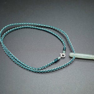 May include: A teal blue braided cord necklace with a light green jade pendant in the shape of a long, thin, rounded rectangle.