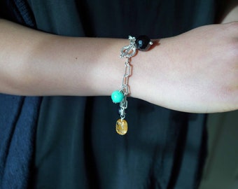 May include: A silver chain bracelet with a turquoise bead, a yellow bead, and a black bead. The bracelet has a decorative charm with a silver design.