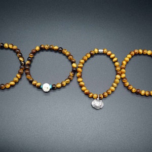 May include: Three tiger's eye beaded bracelets with different charms. The first bracelet has a white bead with a turquoise accent. The second bracelet has a silver charm with a floral design. The third bracelet has no charm.