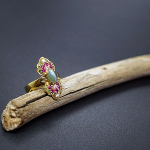 May include: A gold ring with a central oval-shaped, pale green stone, flanked by two ornate, flower-like designs. Each design features red and white gemstones, and is set on a wooden branch against a dark background.