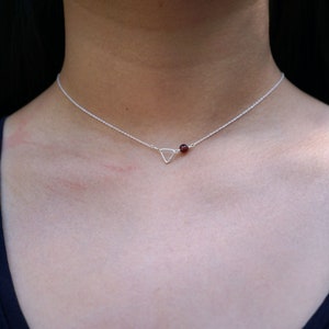 Puede incluir: Un collar de plata con un pequeño colgante triangular y una sola piedra roja.
