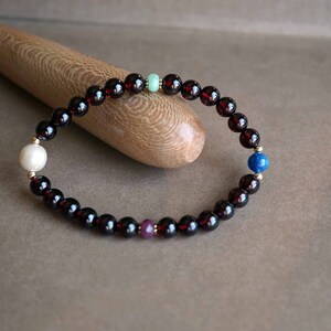 May include: A beaded bracelet featuring dark red garnet beads, interspersed with a white pearl, a light green bead, a blue bead, and a purple bead. The bracelet is strung on a flexible cord and accented with gold-colored beads.