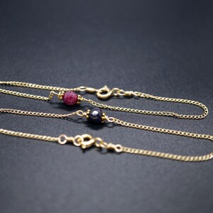 May include: Two delicate gold chain bracelets. One features a red gemstone, the other a dark blue gemstone. Both bracelets have a simple clasp closure. The jewelry is displayed on a dark background.