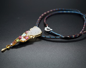 May include: A gold pendant necklace featuring a white stone and colourful gemstones. The necklace is on a brown and blue braided cord.
