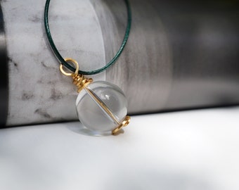 May include: A clear glass sphere pendant necklace with a gold wire wrap and a green cord.