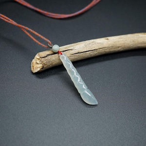 May include: A light blue jade pendant with a carved design, suspended from a red and orange cord. The pendant rests on a piece of driftwood against a dark gray background. The necklace is a unique piece of jewelry.