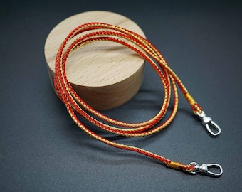 Puede incluir: Un cordón trenzado rojo y dorado con cierres plateados. El cordón está enrollado sobre una superficie de madera.