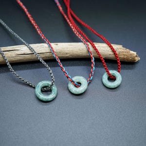Op de afbeelding: Drie kettingen met jade ring hangers op een houten tak. De kettingen zijn gemaakt van gevlochten koorden in grijs, rood en blauw.