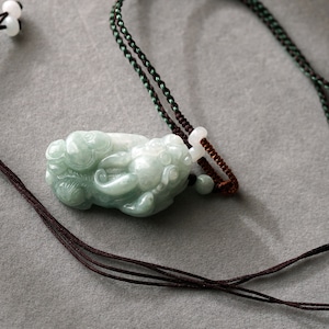 May include: A jade green pendant carved with a detailed design, hanging from a brown and green cord necklace.