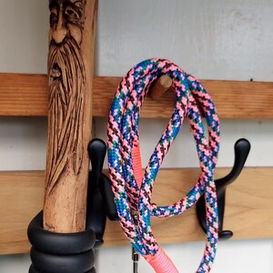 May include: A wooden walking stick with a carved face and a colorful dog lead hanging on a wooden rack. The lead is pink, blue, and brown.