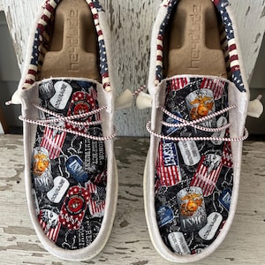 May include: Pair of Hey Dude slip-on shoes with a patriotic theme. The shoes feature a black and gray patterned design with USMC, American flag, and military-themed patches. The shoe trim is white with a red, white, and blue striped pattern.