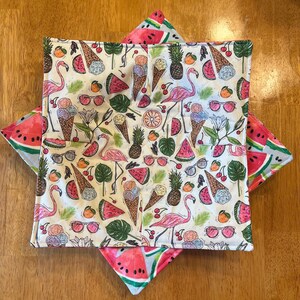 May include: A white, square pot holder with a colorful summer-themed pattern. The pattern includes pink flamingos, watermelon slices, ice cream cones, sunglasses, and pineapples. The pot holder is layered over a watermelon-printed trivet.