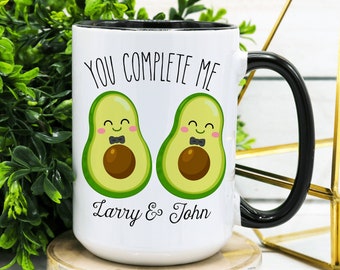 Puede incluir: Taza de cerámica blanca con borde y asa negros. La taza presenta un gráfico de dos mitades de aguacate con caras y pajaritas. El texto "You Complete Me" está por encima de los aguacates y el texto "Larry & John" está por debajo.