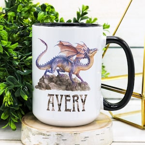 May include: White ceramic mug with a black rim and handle. The mug features a colorful illustration of a dragon with the name "AVERY" printed below.