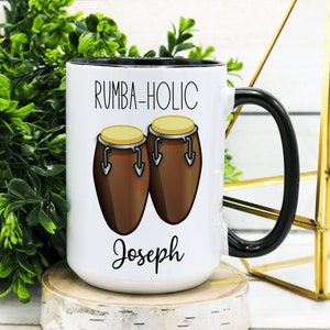 May include: White ceramic mug with a black rim and handle. The mug has a design of two brown conga drums and the text "RUMBA-HOLIC Joseph".