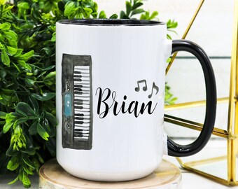 May include: White ceramic mug with a black rim and handle. The mug features a black and white illustration of a keyboard with musical notes and the name "Brian" written in black cursive.
