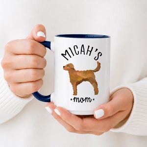 May include: White ceramic mug with a navy blue interior and handle. The mug features the text "Micah's mom" and a brown dog illustration. The mug is held by a person wearing a white sweater.
