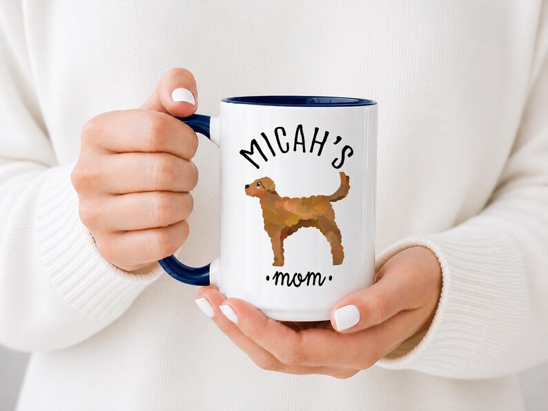 May include: White ceramic mug with a navy blue interior and handle. The mug features the text "Micah's mom" and a brown dog illustration. The mug is held by a person wearing a white sweater.