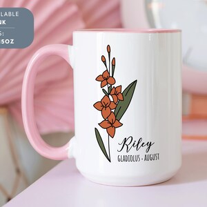 May include: White ceramic mug with a pink rim. The mug features a floral design with orange flowers and green leaves. The text "Riley Gladiolus - August" is printed on the mug.