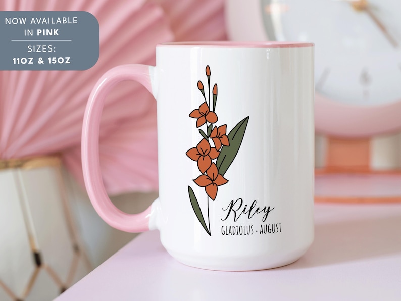 May include: White ceramic mug with a pink rim. The mug features a floral design with orange flowers and green leaves. The text "Riley Gladiolus - August" is printed on the mug.