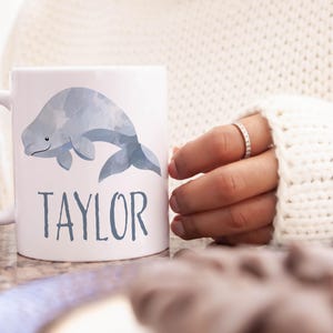 May include: White ceramic mug with a watercolor illustration of a gray beluga whale. The mug has the name "TAYLOR" printed in gray below the whale.