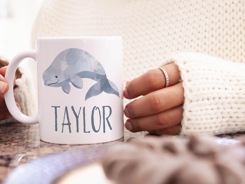 May include: White ceramic mug with a watercolor illustration of a gray beluga whale. The mug has the name "TAYLOR" printed in gray below the whale.