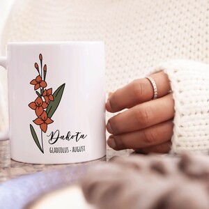 May include: White ceramic mug with a floral design in orange and green. The mug has the name "Dakota" written in cursive with the text "Gladiolus - August" underneath.
