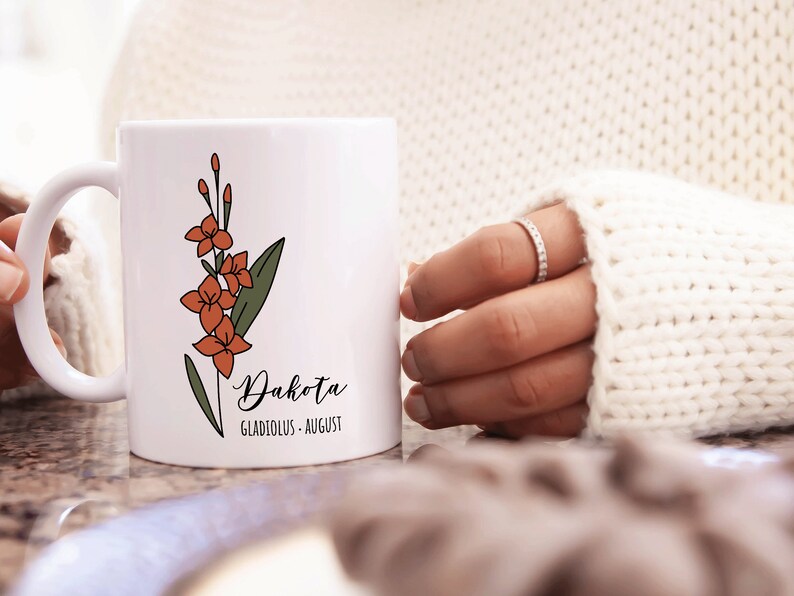 May include: White ceramic mug with a floral design in orange and green. The mug has the name "Dakota" written in cursive with the text "Gladiolus - August" underneath.