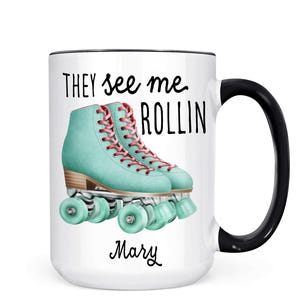 May include: White ceramic coffee mug with a black rim and handle. The mug features a graphic of two teal roller skates with pink laces and the text "They see me rollin'" and the name "Mary" at the bottom.